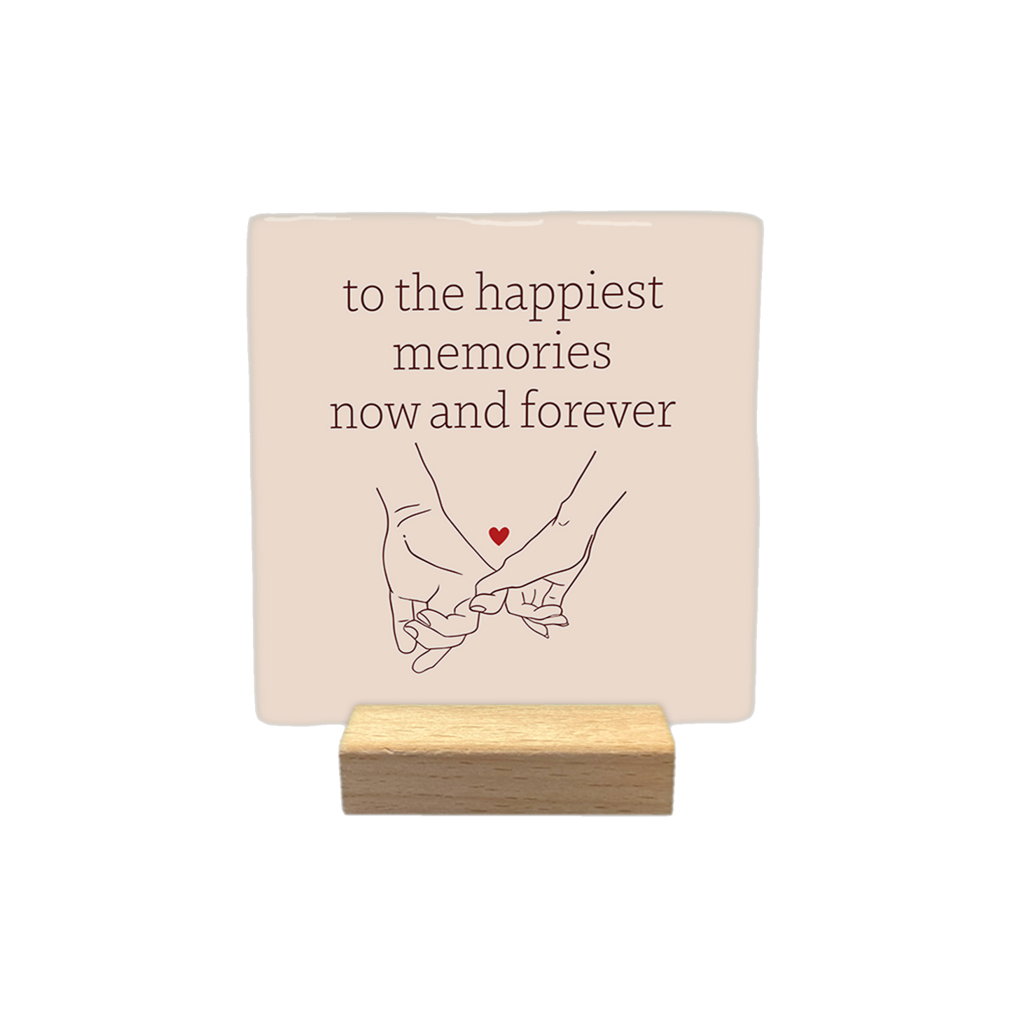 Decoratietegel – 10 x 10 cm -  Op deze decoratietegel staat de tekst "to the happiest memories now and forever" in bruine letters, gecentreerd boven een lijntekening van twee handen die elkaars pink vasthouden. Tussen de handen is een klein, rood hartje afgebeeld. Het ontwerp staat op een lichtbeige achtergrond.