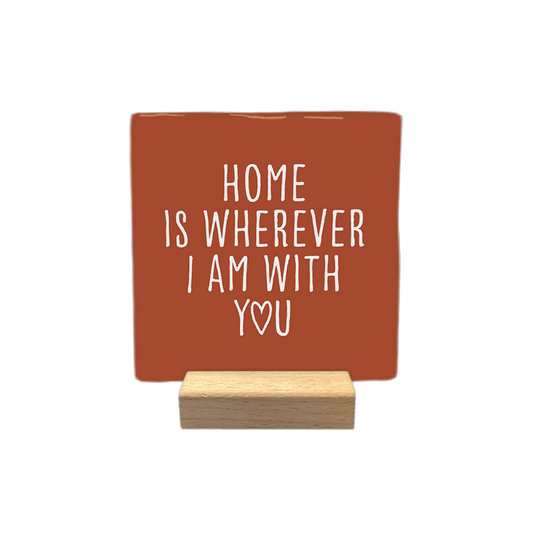 Decoratietegel – 10 x 10 cm - Op deze decoratietegel staat de tekst "HOME IS WHEREVER I AM WITH YOU" in witte letters met een strak, handgeschreven lettertype. De letter "O" in het woord "YOU" is vervangen door een hartje. De tekst is gecentreerd op een roodbruine achtergrond.