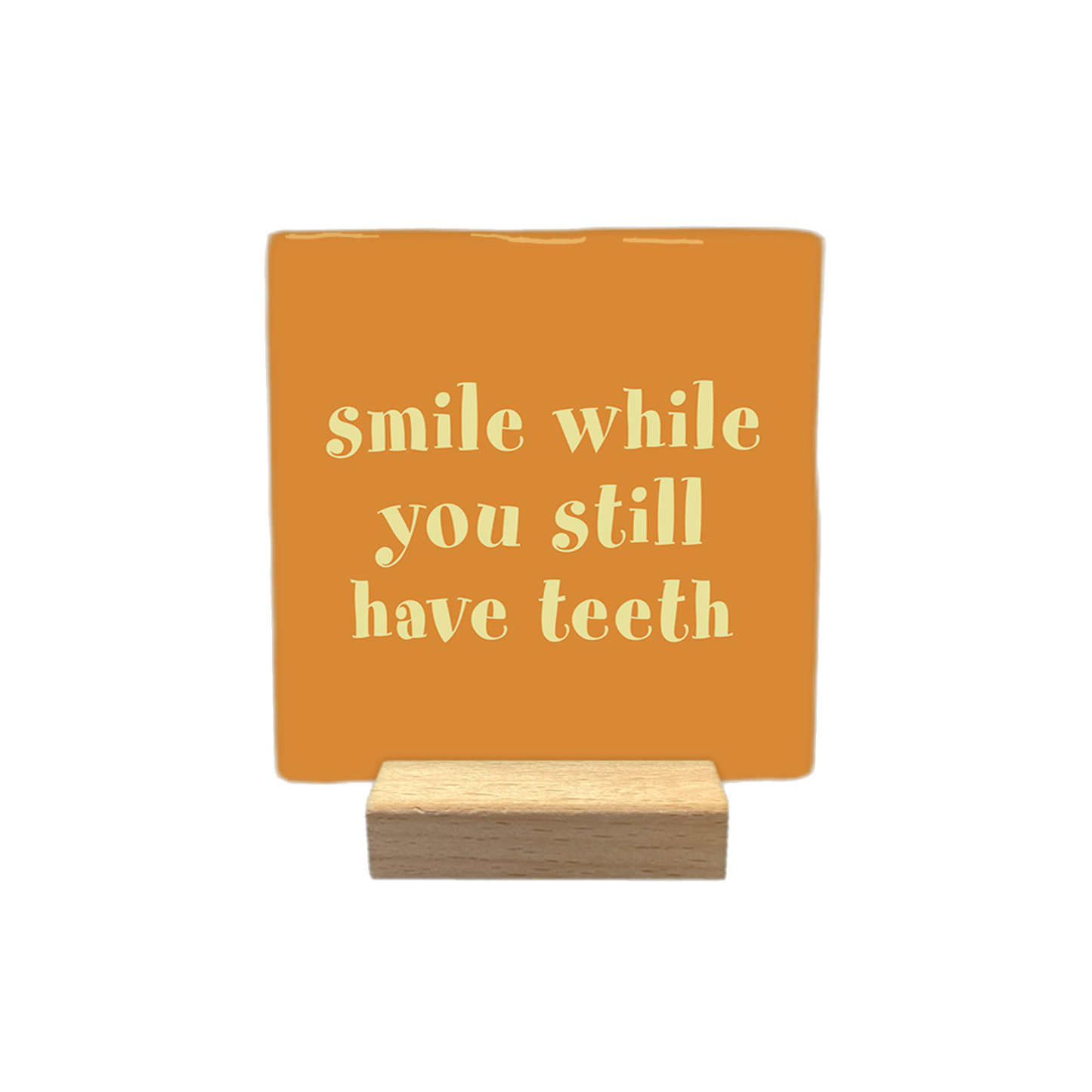 Decoratietegel – 10 x 10 cm - Op deze decoratietegel staat de tekst "smile while you still have teeth" in lichtgele letters. De tekst is gecentreerd op een oranje achtergrond. 