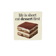 Decoratietegel – 10 x 10 cm - Op deze decoratietegel staat de tekst "life is short eat dessert first" in zwarte letters met een schreeflettertype. Daaronder is een realistische afbeelding van een vierkant stuk tiramisu geplaatst. Het ontwerp is gecentreerd op een crèmekleurige achtergrond. 