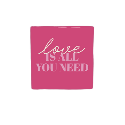 Decoratietegel – 10 x 10 cm - Op deze decoratietegel staat de tekst "love IS ALL YOU NEED" in een combinatie van sierlijke letters voor het woord "love" en strakke hoofdletters voor de rest. De tekst is in lichtroze uitgevoerd en gecentraliseerd op een donkerroze achtergrond. 