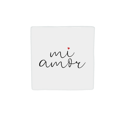  Op deze decoratietegel staat de tekst "mi amor" in een zwart, cursief handschrift. Boven de letter "i" staat een klein, rood hartje. De tekst is gecentreerd op een witte achtergrond. Iedere tegel wordt geleverd inclusief houten presentatieblok. 