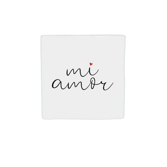  Op deze decoratietegel staat de tekst "mi amor" in een zwart, cursief handschrift. Boven de letter "i" staat een klein, rood hartje. De tekst is gecentreerd op een witte achtergrond. Iedere tegel wordt geleverd inclusief houten presentatieblok. 