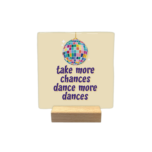 Take more chances dance more dances - Decoratie tegel - Formaat: - Op deze decoratietegel staat een afbeelding van een kleurrijke discobal boven de tekst "take more chances dance more dances" in paarse letters. De tekst en de discobal zijn gecentreerd op een lichtbeige achtergrond. - Snelwenskaart