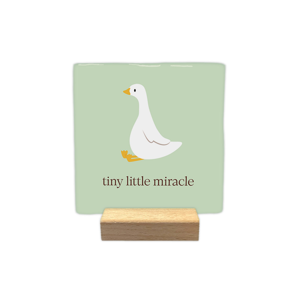 Tiny little miracle - Decoratie tegel - Formaat: 10 x 10 cm - Op deze decoratie tegel staat een gestileerde witte gans met een gele snavel en gele zwemvliezen afgebeeld op een zachtgroene achtergrond. De gans zit in een rustige houding naar links gericht. Onder de illustratie van de vogel staat de tekst "tiny little miracle" geschreven in een bruine kleur. - Snelwenskaart
