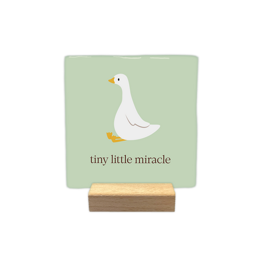 Tiny little miracle - Decoratie tegel - Formaat: 10 x 10 cm - Op deze decoratie tegel staat een gestileerde witte gans met een gele snavel en gele zwemvliezen afgebeeld op een zachtgroene achtergrond. De gans zit in een rustige houding naar links gericht. Onder de illustratie van de vogel staat de tekst "tiny little miracle" geschreven in een bruine kleur. - Snelwenskaart