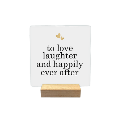 To love laughter and happily ever after - Decoratie tegel - Formaat: - Op deze decoratietegel staat de tekst "to love laughter and happily ever after" in zwarte letters met een elegant schreeflettertype. Bovenaan het ontwerp zijn twee kleine, goudkleurige hartjes afgebeeld. De tekst is gecentreerd op een witte achtergrond. - Snelwenskaart
