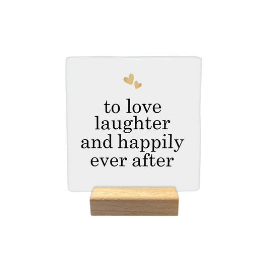 To love laughter and happily ever after - Decoratie tegel - Formaat: - Op deze decoratietegel staat de tekst "to love laughter and happily ever after" in zwarte letters met een elegant schreeflettertype. Bovenaan het ontwerp zijn twee kleine, goudkleurige hartjes afgebeeld. De tekst is gecentreerd op een witte achtergrond. - Snelwenskaart
