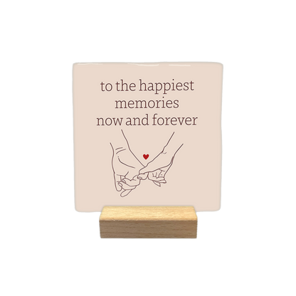 To the happiest memories now and forever - Decoratie tegel - Formaat: - Op deze decoratietegel staat de tekst "to the happiest memories now and forever" in bruine letters, gecentreerd boven een lijntekening van twee handen die elkaars pink vasthouden. Tussen de handen is een klein, rood hartje afgebeeld. Het ontwerp staat op een lichtbeige achtergrond. - Snelwenskaart