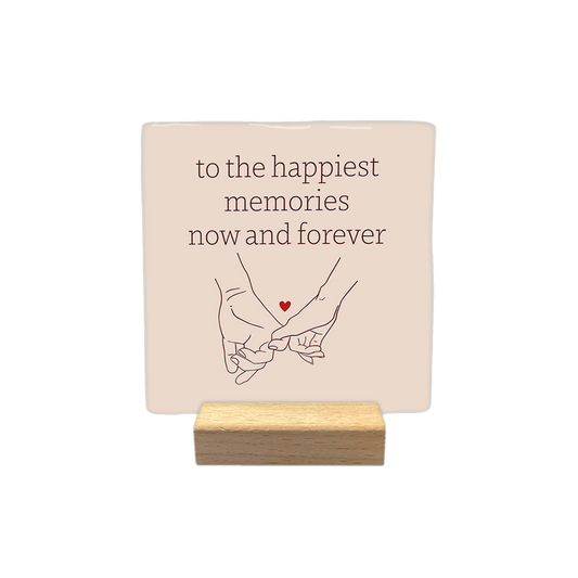 To the happiest memories now and forever - Decoratie tegel - Formaat: - Op deze decoratietegel staat de tekst "to the happiest memories now and forever" in bruine letters, gecentreerd boven een lijntekening van twee handen die elkaars pink vasthouden. Tussen de handen is een klein, rood hartje afgebeeld. Het ontwerp staat op een lichtbeige achtergrond. - Snelwenskaart