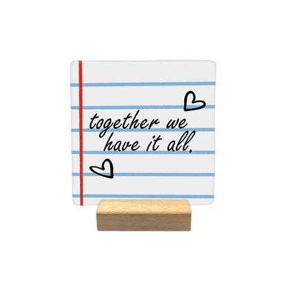 Together we have it all - Decoratie tegel - Formaat: - Op deze decoratietegel staat de tekst "together we have it all." in een zwart, cursief lettertype. De achtergrond heeft het uiterlijk van gelinieerd schriftpapier met blauwe horizontale lijnen en een rode verticale kantlijn. Het ontwerp wordt aangevuld met twee handgetekende zwarte hartjes. - Snelwenskaart