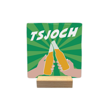 Tsjoch - Decoratie tegel - Formaat: - Op deze decoratie tegel staat de tekst "TSJOCH" in witte letters bovenaan afgebeeld. Onder de tekst is een afbeelding te zien van twee handen die een toost uitbrengen met bierflesjes. De achtergrond heeft een groenkleurig patroon met stralen. - Snelwenskaart