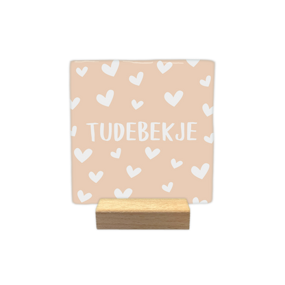 Tudebekje - Decoratie tegel - Formaat: - Op deze decoratie tegel staat de tekst "tudebekje" in witte letters centraal afgebeeld. De achtergrond van de tegel is lichtbeige van kleur en is versierd met een patroon van kleine witte hartjes. - Snelwenskaart