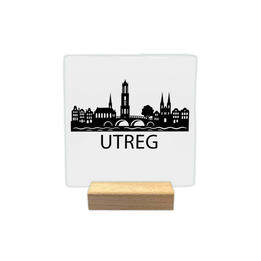 Utreg skyline - Decoratie tegel - Formaat: - Op deze decoratie tegel staat een zwarte skyline van Utrecht afgebeeld tegen een witte achtergrond. Het silhouet bevat een brug over het water, de Domtoren en historische panden. Onder de skyline staat in zwarte letters de tekst "Utreg". Het ontwerp is minimalistisch uitgevoerd in zwart en wit. - Snelwenskaart