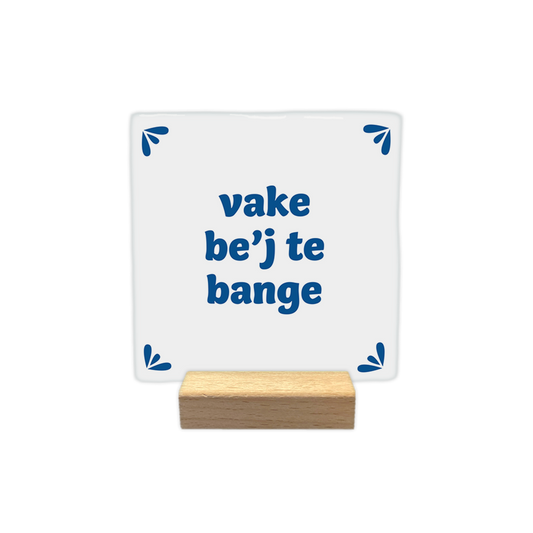 Vake be'j te bange - Decoratie tegel - Formaat: - Op deze decoratie tegel staat de tekst "vake be’j te bange" in blauwe letters centraal afgebeeld. In elk van de vier hoeken is een klein, blauw decoratief element geplaatst dat uit drie bladvormige blaadjes bestaat. De achtergrond van de tegel is wit. - Snelwenskaart