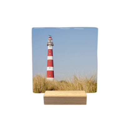 Vuurtoren Ameland - Decoratie tegel - Formaat: - Op deze decoratie tegel is een vuurtoren met rood-witte horizontale banden afgebeeld. De vuurtoren staat achter een voorgrond van duingras. De lucht op de achtergrond is helderblauw. - Snelwenskaart