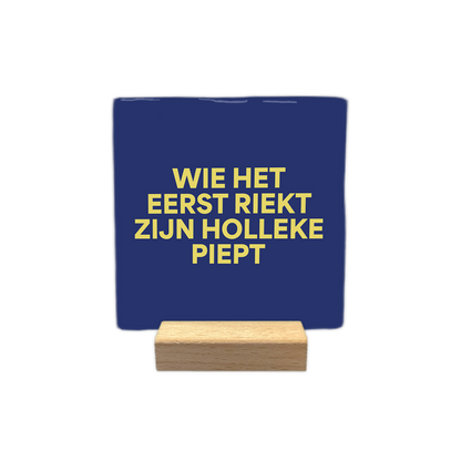 Wie het eerst riekt zien Holleke piept - Decoratie tegel - Formaat: - Op deze decoratie tegel staat de tekst "wie het eerst riekt zijn holleke piept" in vier regels afgebeeld. De tekst is geschreven in gele letters. De achtergrond van de tegel is donkerblauw. - Snelwenskaart