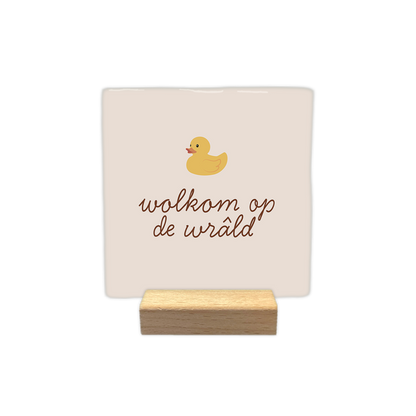Wolkom op de wrâld - Decoratie tegel - Formaat: - Op deze decoratie tegel staat een illustratie van een geel badeendje afgebeeld. Onder het eendje staat in bruine, sierlijke letters de tekst "wolkom op de wrâld". De achtergrond van de tegel heeft een zachte, lichtbeige kleur. - Snelwenskaart