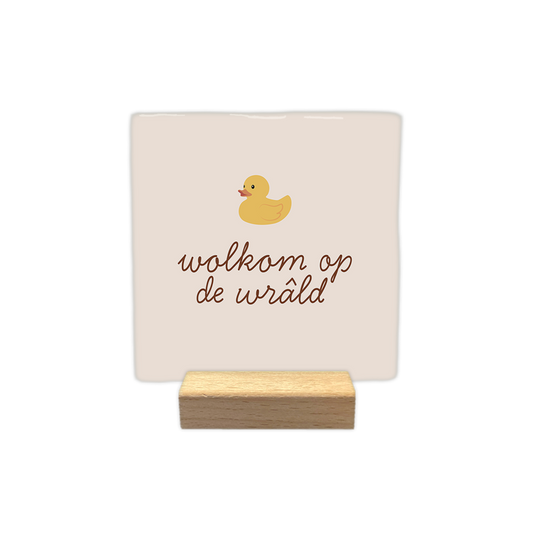 Wolkom op de wrâld - Decoratie tegel - Formaat: - Op deze decoratie tegel staat een illustratie van een geel badeendje afgebeeld. Onder het eendje staat in bruine, sierlijke letters de tekst "wolkom op de wrâld". De achtergrond van de tegel heeft een zachte, lichtbeige kleur. - Snelwenskaart