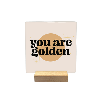 You are golden - Decoratie tegel - Formaat: - Op deze decoratietegel staat de tekst "you are golden" in zwarte letters met een afgerond, speels lettertype. De tekst is gecentreerd over een goudkleurige cirkel en wordt omringd door kleine, gestileerde sterretjes. Het gehele ontwerp bevindt zich op een crèmekleurige achtergrond. - Snelwenskaart