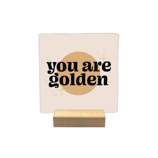 You are golden - Decoratie tegel - Formaat: - Op deze decoratietegel staat de tekst "you are golden" in zwarte letters met een afgerond, speels lettertype. De tekst is gecentreerd over een goudkleurige cirkel en wordt omringd door kleine, gestileerde sterretjes. Het gehele ontwerp bevindt zich op een crèmekleurige achtergrond. - Snelwenskaart