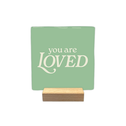 You are loved - Decoratie tegel - Formaat: - Op deze decoratie tegel staat de tekst "you are loved" centraal in crèmekleurige letters. Het woord "loved" is in een groter, opvallend lettertype geschreven, terwijl "you are" erboven in een kleiner lettertype staat. Het geheel is geplaatst tegen een zachtgroene achtergrond. - Snelwenskaart