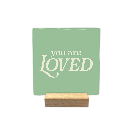 You are loved - Decoratie tegel - Formaat: - Op deze decoratie tegel staat de tekst "you are loved" centraal in crèmekleurige letters. Het woord "loved" is in een groter, opvallend lettertype geschreven, terwijl "you are" erboven in een kleiner lettertype staat. Het geheel is geplaatst tegen een zachtgroene achtergrond. - Snelwenskaart