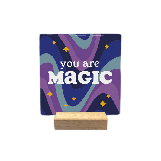 You are magic - Decoratie tegel - Formaat: - Op deze decoratietegel staat de tekst "you are MAGIC" centraal afgebeeld, waarbij "you are" in een kleiner, wit lettertype boven het grotere, opvallende witte woord "MAGIC" staat. De achtergrond bestaat uit golvende banen in tinten blauw en paars, wat een dromerig effect geeft. De illustratie is versierd met kleine, gele sterretjes die over de achtergrond verspreid staan. - Snelwenskaart