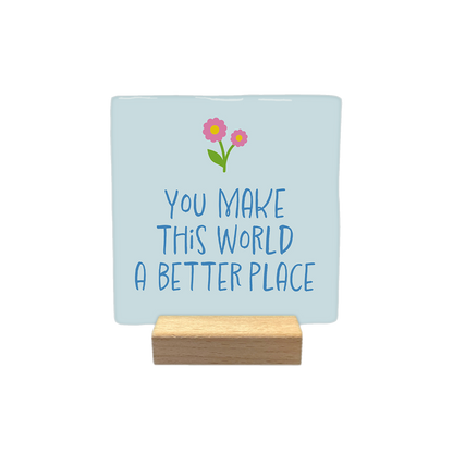 You make this world a better place - Decoratie tegel - Formaat: - Op deze decoratieteel staat de tekst 'You make this world a better place' geschreven in donderblauwe letters. Boven de tekst staan twee bloeken in de kleur roze met een gele knop en een groen blaadje. De tekst staat gecentreerd op een lichtblauwe achtergrond. - Snelwenskaart