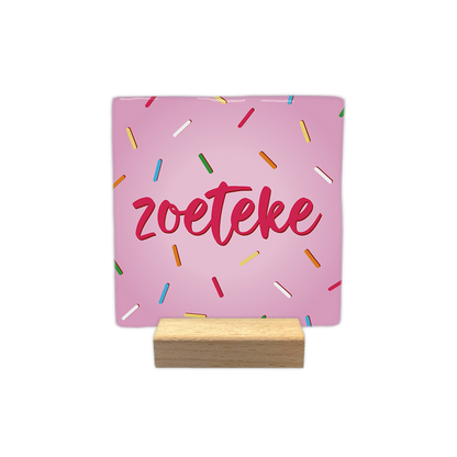 Zoeteke - Decoratie tegel - Formaat: - Op deze decoratie tegel staat het woord "zoeteke" centraal afgebeeld in roze, handgeschreven letters. De achtergrond van de tegel is lichtroze en is versierd met een patroon van kleine, gekleurde staafjes die doen denken aan spikkels. - Snelwenskaart