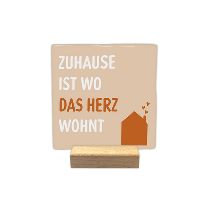 Zuhause ist wo das herz wohnt - Decoratie tegel - Formaat: - Op deze decoratie tegel staat de tekst "zuhause ist wo das herz wohnt" afgebeeld. De tekst is verdeeld over vier regels, waarbij de woorden "zuhause", "ist" en "wohnt" in het wit zijn geschreven en het woord "herz" in het bruin. Rechtsonder staat een bruine illustratie van een huis met drie kleine hartjes die uit de schoorsteen stijgen. De achtergrond van het ontwerp is lichtbeige. - Snelwenskaart
