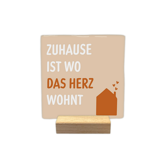 Zuhause ist wo das herz wohnt - Decoratie tegel - Formaat: - Op deze decoratie tegel staat de tekst "zuhause ist wo das herz wohnt" afgebeeld. De tekst is verdeeld over vier regels, waarbij de woorden "zuhause", "ist" en "wohnt" in het wit zijn geschreven en het woord "herz" in het bruin. Rechtsonder staat een bruine illustratie van een huis met drie kleine hartjes die uit de schoorsteen stijgen. De achtergrond van het ontwerp is lichtbeige. - Snelwenskaart