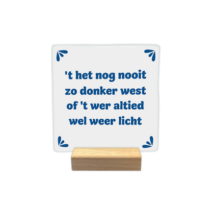 t het nog nooit zo donker west - Decoratie tegel - Formaat: - Op deze decoratie tegel staat de tekst "'t het nog nooit zo donker west of 't wer altied wel weer licht" in blauwe letters centraal afgebeeld. De achtergrond van de tegel is wit. In elke hoek van de tegel is een klein, blauw decoratief motiefje geplaatst. - Snelwenskaart