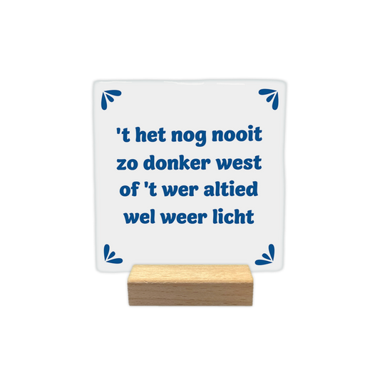 t het nog nooit zo donker west - Decoratie tegel - Formaat: - Op deze decoratie tegel staat de tekst "'t het nog nooit zo donker west of 't wer altied wel weer licht" in blauwe letters centraal afgebeeld. De achtergrond van de tegel is wit. In elke hoek van de tegel is een klein, blauw decoratief motiefje geplaatst. - Snelwenskaart